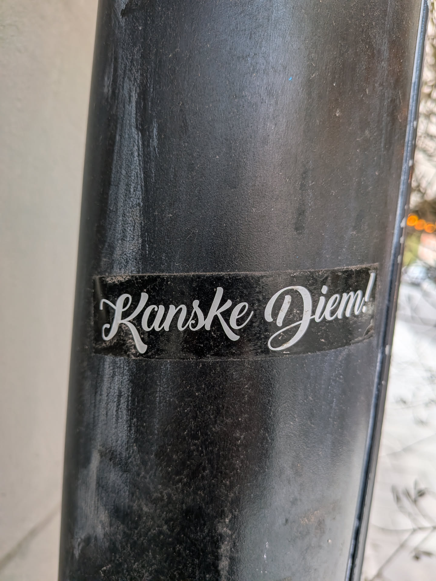 A rectangular sticker featuring white cursive text on a solid black background. The sticker is applied to a textured, dark grey or black pole and shows signs of dust and minor wear, particularly around its edges. The text 'Kanske Diem!' is centrally placed and rendered in an elegant, flowing script, suggesting a humorous or ironic take on 'Carpe Diem'.