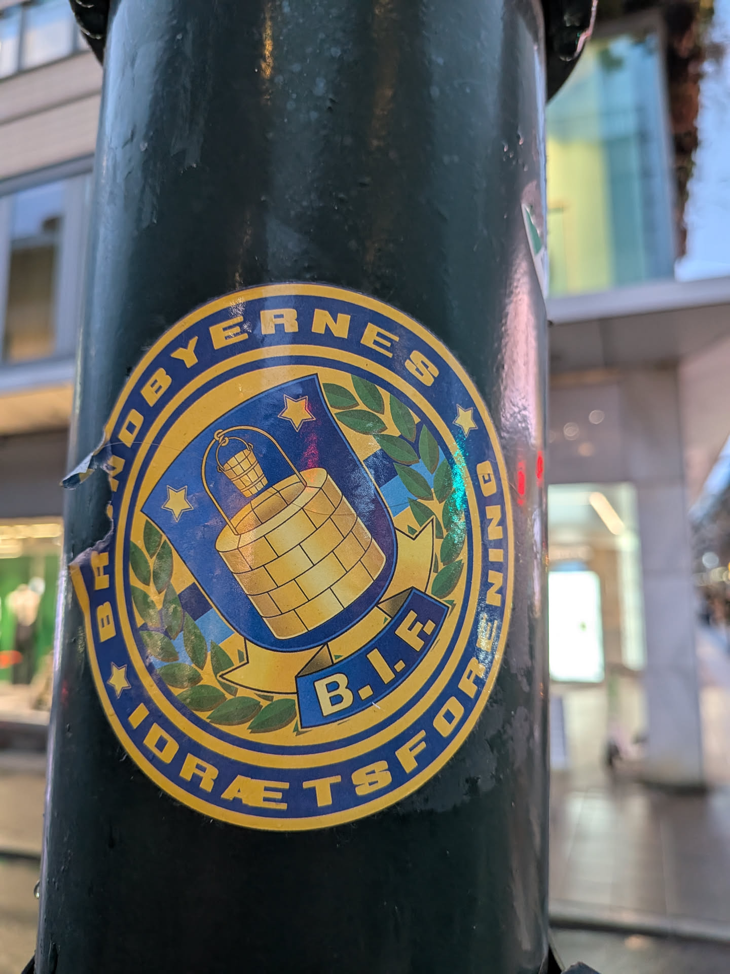 The sticker is circular with a yellow border. Inside, a blue shield is prominent, depicting a golden well with a bucket hanging from a rope. Laurel branches flank the shield. The text 'BRØDBYERNES B.I.F. IDRÆTSFORBUND' encircles the shield in yellow lettering on a blue background. Two small yellow stars are also visible within the shield. The sticker appears to be slightly worn, with some peeling at the edges, suggesting it has been affixed for some time.
