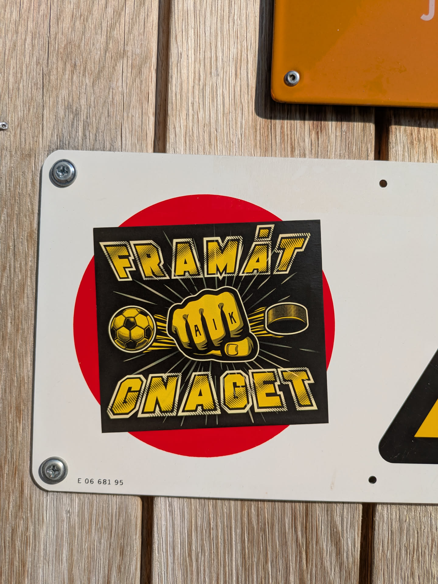 The sticker depicts a black square with a red circle behind it. In yellow text, the words "FRAMÅT" appear at the top and "GNAGET" at the bottom. In the center, there is a yellow fist clenched with the letters "AIK" visible on the knuckles, a soccer ball on the left side of the fist, and a hockey puck on the right.