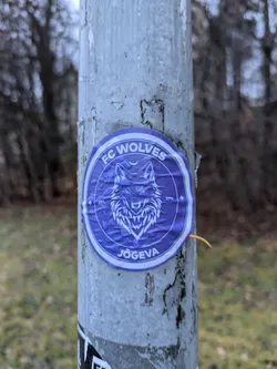 Street sticker A purple circular sticker with a wolf's head in the center. The text "FC Wolves" is at the top, and "Jõgeva" at the bottom.  There are small symbols on either side of the wolf's head, and the year 2016 is visible.