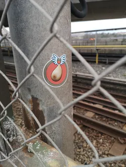 The sticker is circular, featuring a prominent white outer border. Inside, there's a thin black line, followed by a light grey ring, a darker grey-brown ring, and an inner red-orange circle. At the center of this red-orange circle is a stylized, light-skinned breast with a brown nipple. Attached to the top of the breast are two cartoon-style wings, predominantly white with light blue outlines and feather details, giving the impression of flight.