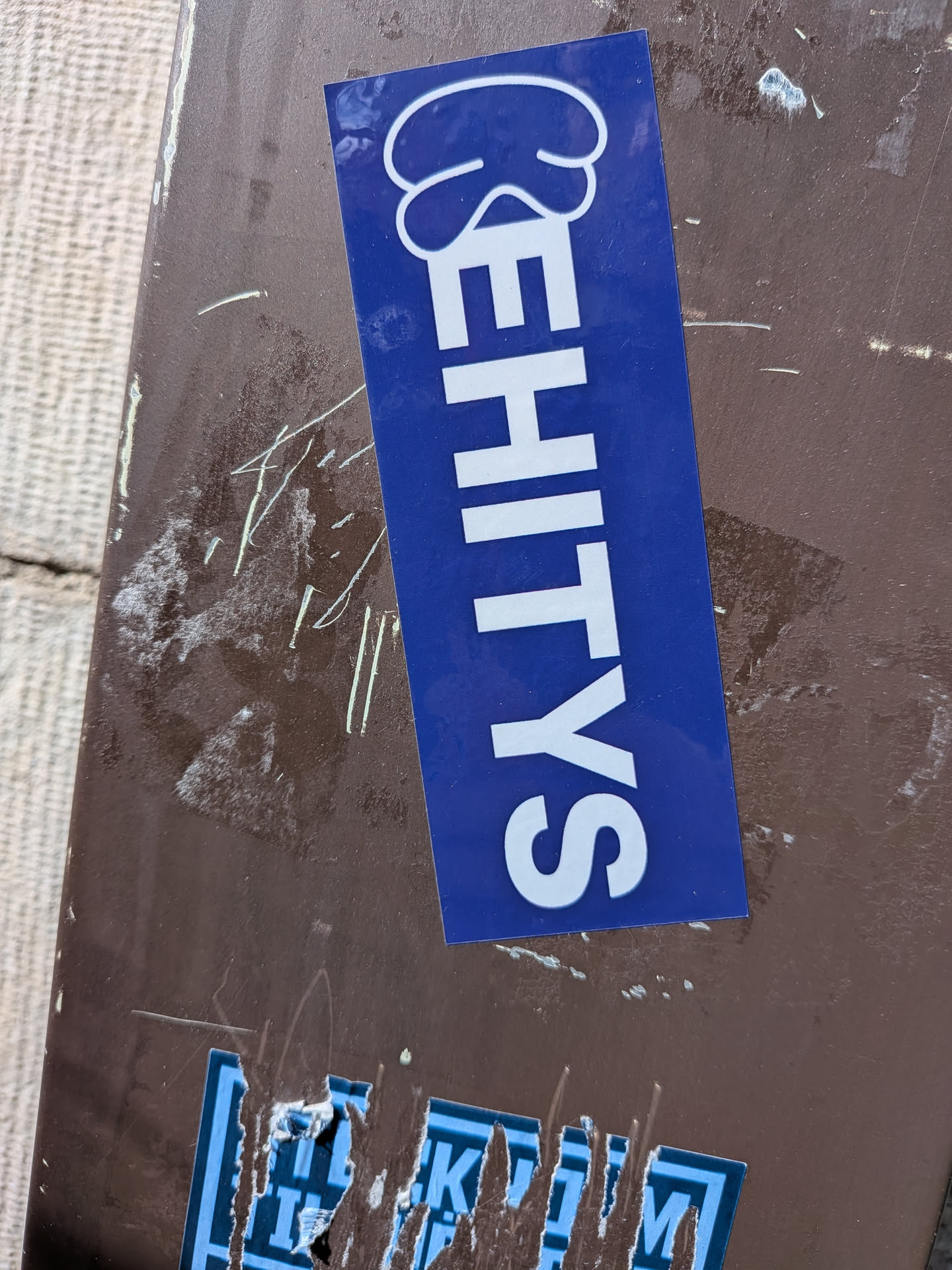 A rectangular sticker with a vibrant blue background, oriented vertically. The word "EHITYS" is written in bold, sans-serif, uppercase white letters, stacked from bottom to top. Above the 'E' (at the top of the sticker), there is a white, stylized graphic resembling a cloud or a thought bubble. The sticker has a glossy finish and is applied to a dark brown, textured surface.