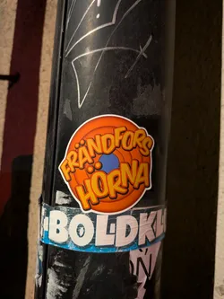 The sticker is circular with a clean white border. Its background features concentric rings of orange, transitioning from a lighter orange in the center to a darker, almost reddish-orange at the edges, strongly reminiscent of the classic Looney Tunes logo. In the very center, there is a small, solid blue circle. The text 'FRÅNDFORS HORNA' is prominently displayed in two lines, with 'FRÅNDFORS' on top and 'HORNA' below. The letters are bold, cartoonish, and yellow with a thick orange outline and a subtle darker orange shadow, giving them a playful, three-dimensional effect. The sticker is affixed to a dark, grimy utility pole, contrasting sharply with its bright colors.