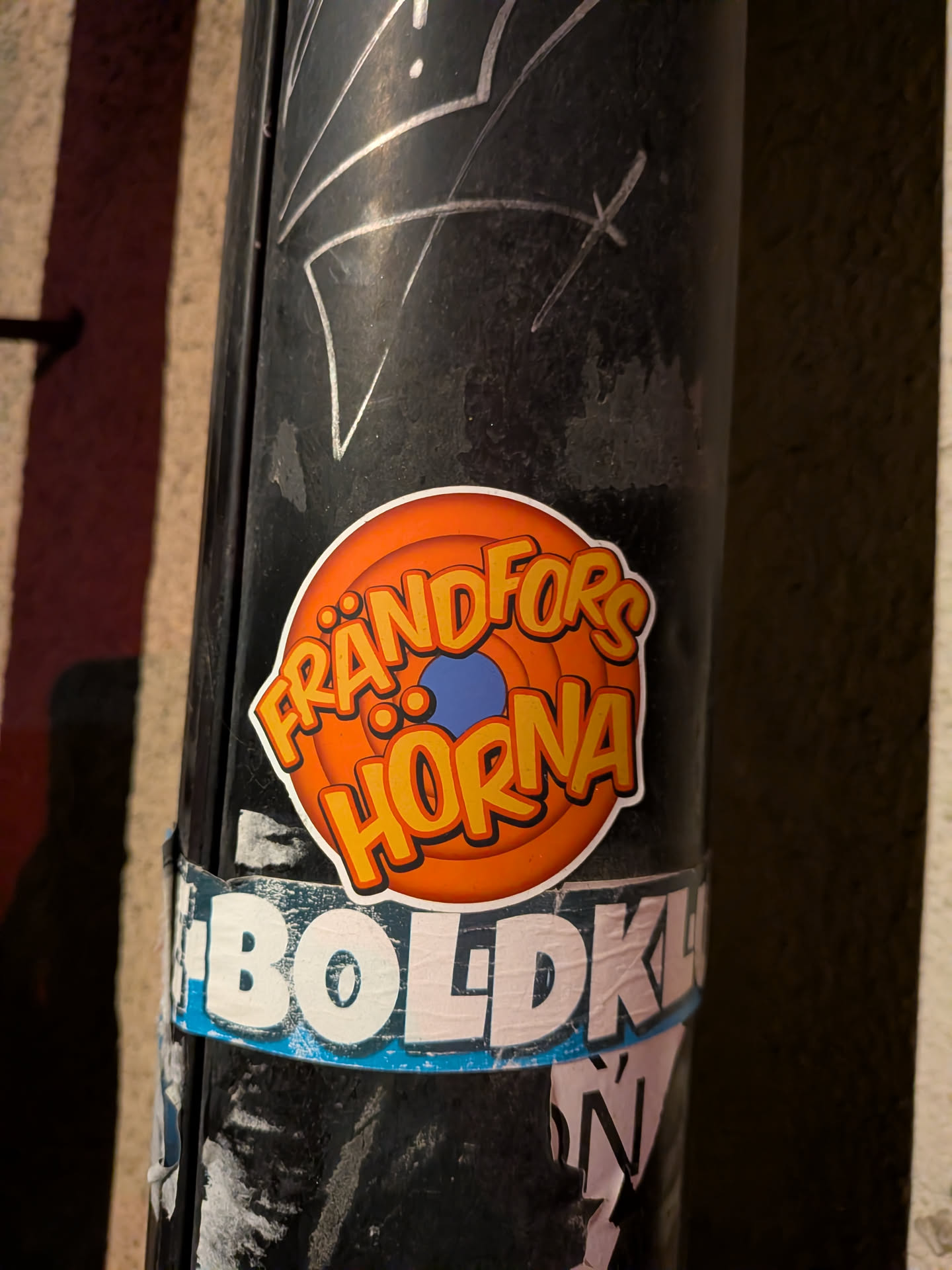 The sticker is circular with a clean white border. Its background features concentric rings of orange, transitioning from a lighter orange in the center to a darker, almost reddish-orange at the edges, strongly reminiscent of the classic Looney Tunes logo. In the very center, there is a small, solid blue circle. The text 'FRÅNDFORS HORNA' is prominently displayed in two lines, with 'FRÅNDFORS' on top and 'HORNA' below. The letters are bold, cartoonish, and yellow with a thick orange outline and a subtle darker orange shadow, giving them a playful, three-dimensional effect. The sticker is affixed to a dark, grimy utility pole, contrasting sharply with its bright colors.