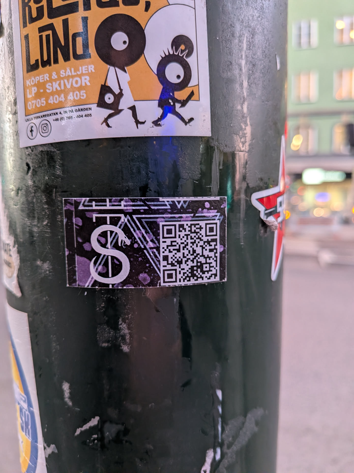 Street sticker The sticker is rectangular with a dark, speckled background, predominantly black and purple. On the left side, a stylized white symbol resembling a snake coiled around a vertical line is prominently displayed. To the right of the symbol is a standard QR code. The edges of the sticker appear slightly worn.