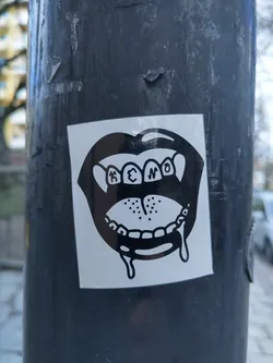 Street sticker A black and white sticker depicting a cartoonish mouth with teeth dripping. The text 'K3N0' is visible within the mouth.  The sticker is affixed to a dark grey pole.