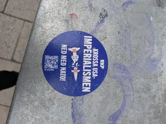 Street sticker A circular blue sticker affixed to a metallic surface. The sticker features a faint outline of a world map in the background. In the center, there is a graphic of a white missile broken in half; the top half is marked with dollar signs ($) and red stripes. To the right of the graphic, vertical white text reads 'KROSSA USA-IMPERIALISMEN' (Crush US Imperialism) below a small logo for 'RKP'. To the left, vertical white text reads 'NED MED NATO!' (Down with NATO!). On the far left, there is a QR code next to the website 'marxist.se'.