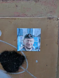 Street sticker The sticker features a close-up portrait of a smiling man wearing a dark military-style cap with a Swedish emblem. The background is blurred and appears to be an urban setting with blue tones. White text is overlaid on the lower part of the image. The sticker itself is square with slightly rounded corners.