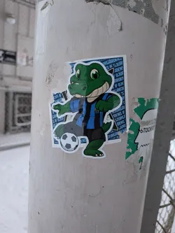 Street sticker The sticker features a cartoonish green alligator or crocodile mascot with large green eyes and a wide, friendly smile. It is wearing a blue and black striped short-sleeved jersey and dark grey shorts. Its left foot is positioned on a classic black and white soccer ball. The background of the sticker is a repeating pattern of white text that reads 'DJURGÅRDENS IF' on a blue field. The entire character has a clean white outline, making it stand out. The sticker is affixed to a light-colored pole, showing some signs of wear and tear around its edges.
