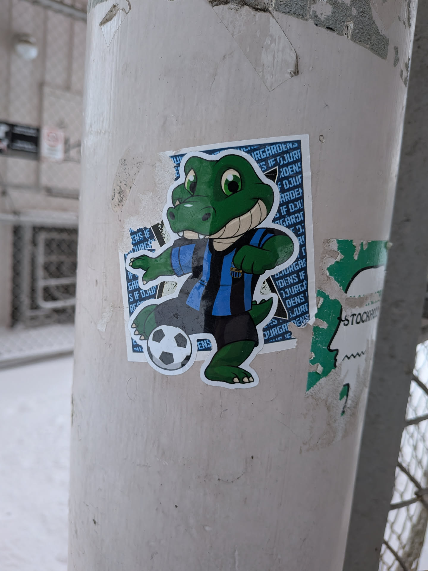 Street sticker The sticker features a cartoonish green alligator or crocodile mascot with large green eyes and a wide, friendly smile. It is wearing a blue and black striped short-sleeved jersey and dark grey shorts. Its left foot is positioned on a classic black and white soccer ball. The background of the sticker is a repeating pattern of white text that reads 'DJURGÅRDENS IF' on a blue field. The entire character has a clean white outline, making it stand out. The sticker is affixed to a light-colored pole, showing some signs of wear and tear around its edges.