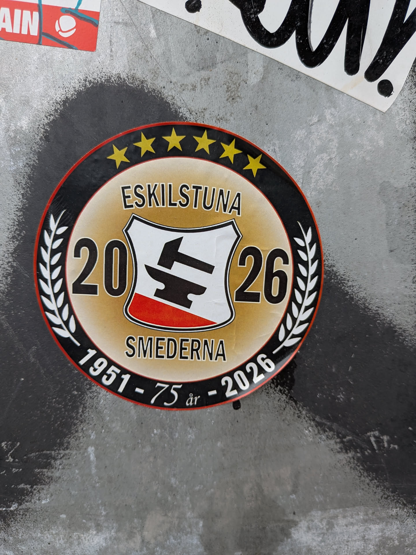 A circular sticker celebrating the 75th anniversary of the Eskilstuna Smederna speedway club. It features a central shield with a black hammer and anvil logo, flanked by the year '2026'. The outer black ring contains five yellow stars, white laurel wreaths, and the text '1951 - 75 år - 2026'.