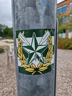 A sticker featuring a green and gold emblem with a star, oars, and laurel wreath. The sticker appears to be affixed to a metal pole outdoors.  There is some minor damage to the sticker.