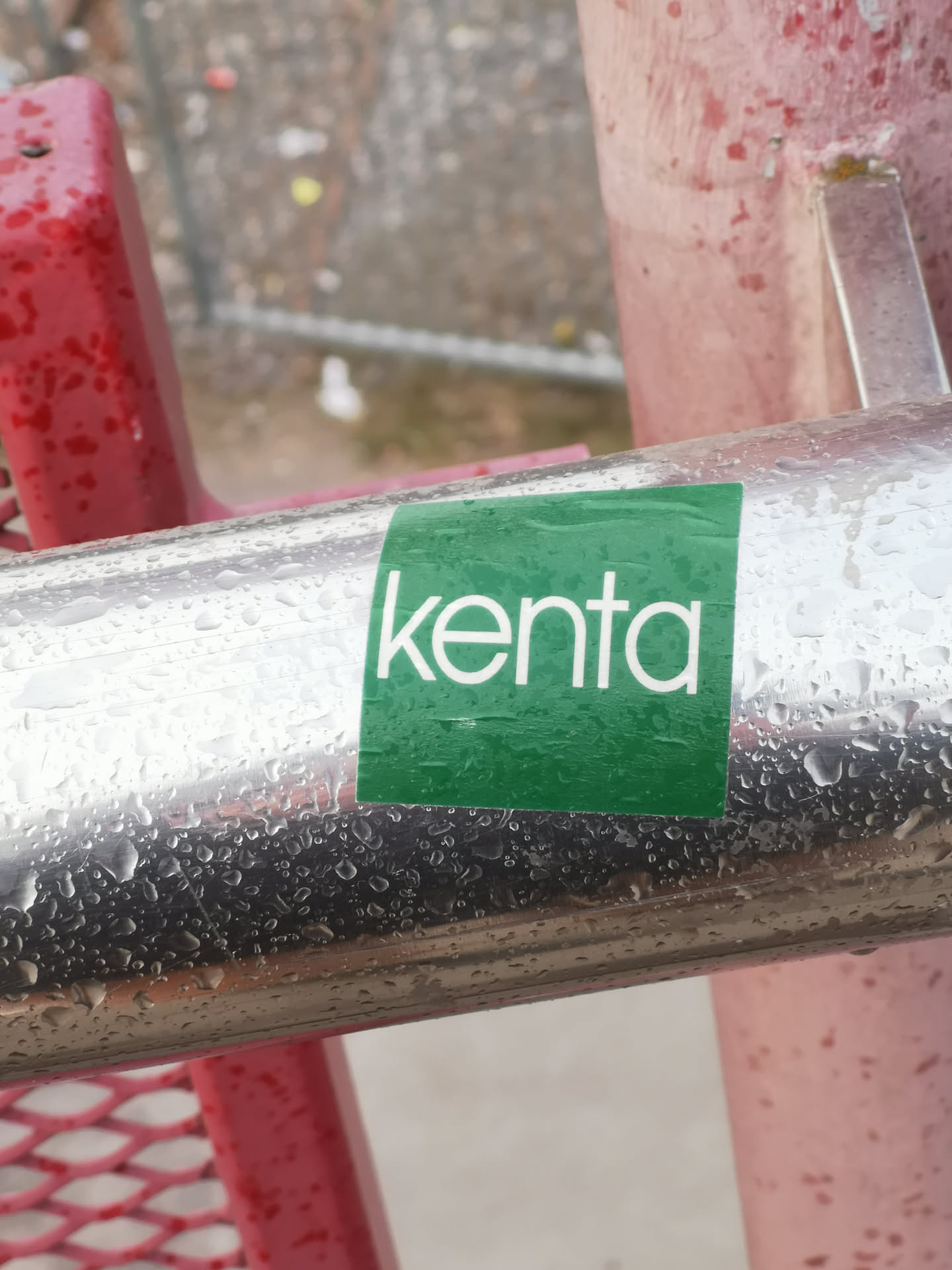 A rectangular sticker with a vibrant green background, featuring the word 'kenta' in clean, white, lowercase sans-serif letters. The sticker appears slightly wrinkled and is covered in numerous small water droplets, indicating it is wet. It is affixed to a reflective, chrome-like metal pole, also covered in water droplets.