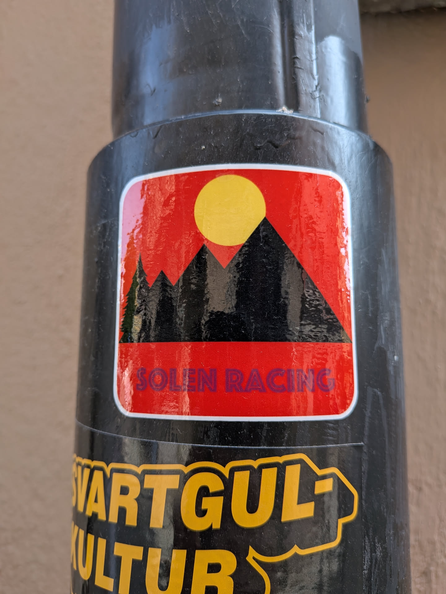 A rounded rectangular sticker featuring black mountain silhouettes, a single pine tree, and a yellow sun against a red background. The text 'SOLEN RACING' is printed in purple at the bottom.