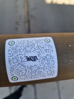 Street sticker A white square sticker with a QR code and some graffiti-style text in the center. The sticker is affixed to a brown cylindrical object outdoors.  The QR code appears to be a standard QR code.
