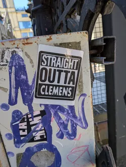 Street sticker The sticker is a white rectangle with slightly rounded corners, featuring a prominent black border. Inside the border, the background is black, and the text "STRAIGHT OUTTA CLEMENS" is displayed in bold, white, sans-serif, uppercase letters. The words are stacked vertically, with "STRAIGHT" on the top line, "OUTTA" on the middle line, and "CLEMENS" on the bottom line, each centered. The sticker is adhered to a light-colored, rusty metal surface, partially overlapping purple paint or graffiti.