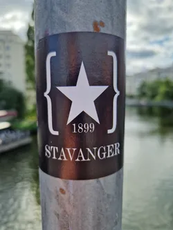Street sticker A dark-colored sticker with a white star and the year 1899, along with the word 'Stavanger' printed in a serif typeface.  The star is enclosed in stylized curly brackets. The sticker is affixed to a metal pole by a body of water.
