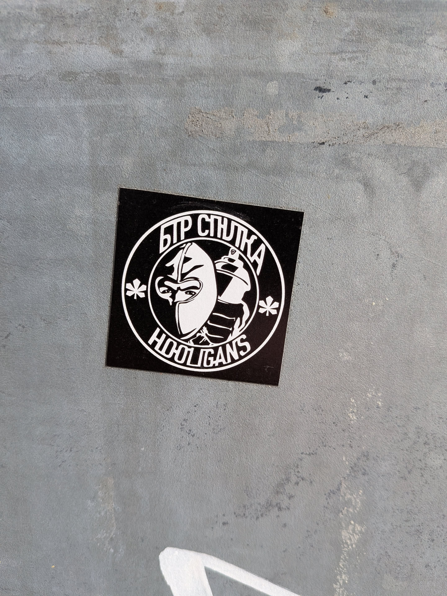 The sticker is square-shaped with a black background and white graphics. Centered on the sticker is a circular emblem. Within the circle, a stylized mask with sharp lines covers the face of a figure, with visible eyes peering out. To the right of the mask, a spray can is depicted, also in white. The text 'БТР СПУТНИКА' is written in a curved line above the mask, and 'HOOLIGANS' is written below it, also in a curve. Two small, asterisk-like shapes are positioned to the left and right of the mask within the circle. The overall design is bold and has a graphic, urban aesthetic.
