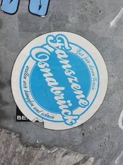 Street sticker A sticker featuring the text "Fanszene Cosmaulbrück" in a stylized font. The text is written in German and translates to "Fan scene Cosmaulbrück". The sticker is circular with a blue central area and a white outer ring. There is additional text in the outer ring, which appears to be a motto or slogan.