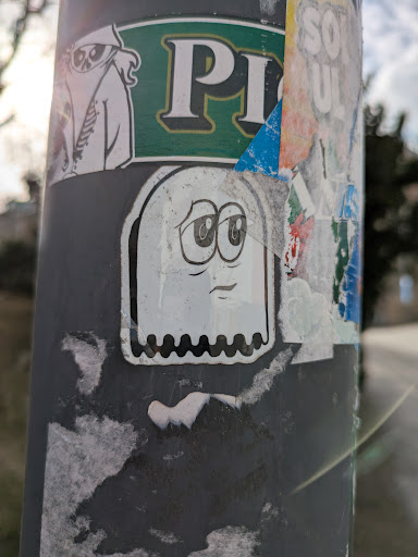 A white, cartoonish ghost sticker with large, sad eyes and a downturned mouth is stuck to a pole. The ghost has a rectangular shape with a wavy bottom edge. The background consists of other worn and partially torn stickers, adding to the urban texture.