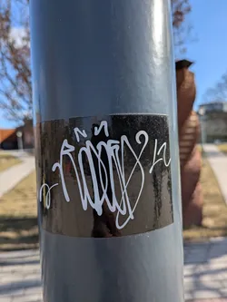 The sticker is a rectangular, horizontally oriented piece with a glossy black background. Prominently displayed on this black surface is a highly stylized, flowing graffiti tag rendered in thick, opaque white lines. The tag features intricate loops, curves, and sharp angles, characteristic of a complex handstyle, making the individual letters largely illegible. Elements that could be interpreted as an 'N' or 'M' are visible at the top left, and a '2' or 'L' shape appears towards the right. The white lines are crisp against the dark background, and the sticker shows minor reflections from the surrounding environment.