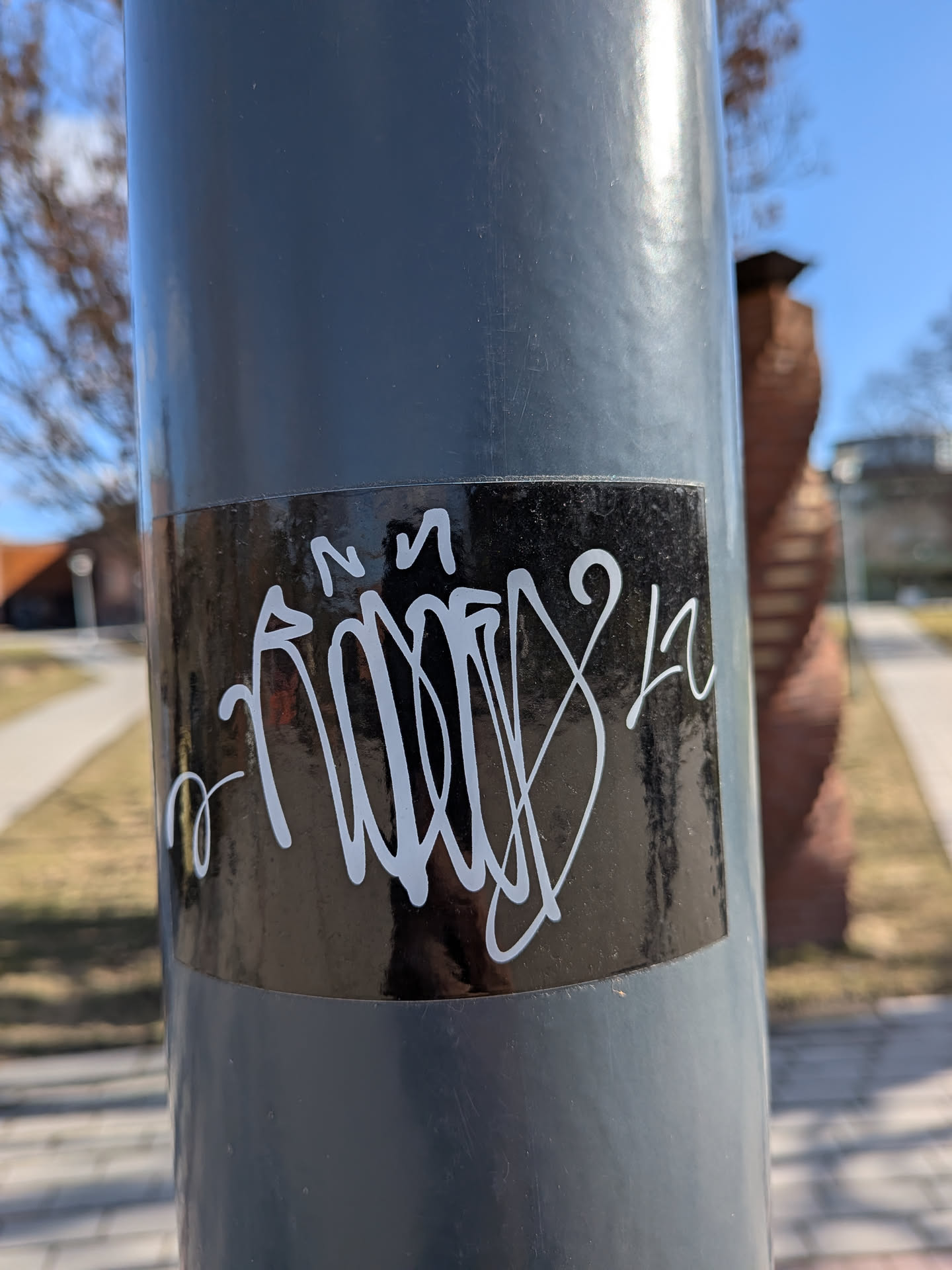 The sticker is a rectangular, horizontally oriented piece with a glossy black background. Prominently displayed on this black surface is a highly stylized, flowing graffiti tag rendered in thick, opaque white lines. The tag features intricate loops, curves, and sharp angles, characteristic of a complex handstyle, making the individual letters largely illegible. Elements that could be interpreted as an 'N' or 'M' are visible at the top left, and a '2' or 'L' shape appears towards the right. The white lines are crisp against the dark background, and the sticker shows minor reflections from the surrounding environment.