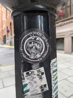 Street sticker A circular black and white sticker depicting a character in a gas mask and helmet, surrounded by a circular border containing the text 'zinloos geluid', 'high risk', and 'low bass'.