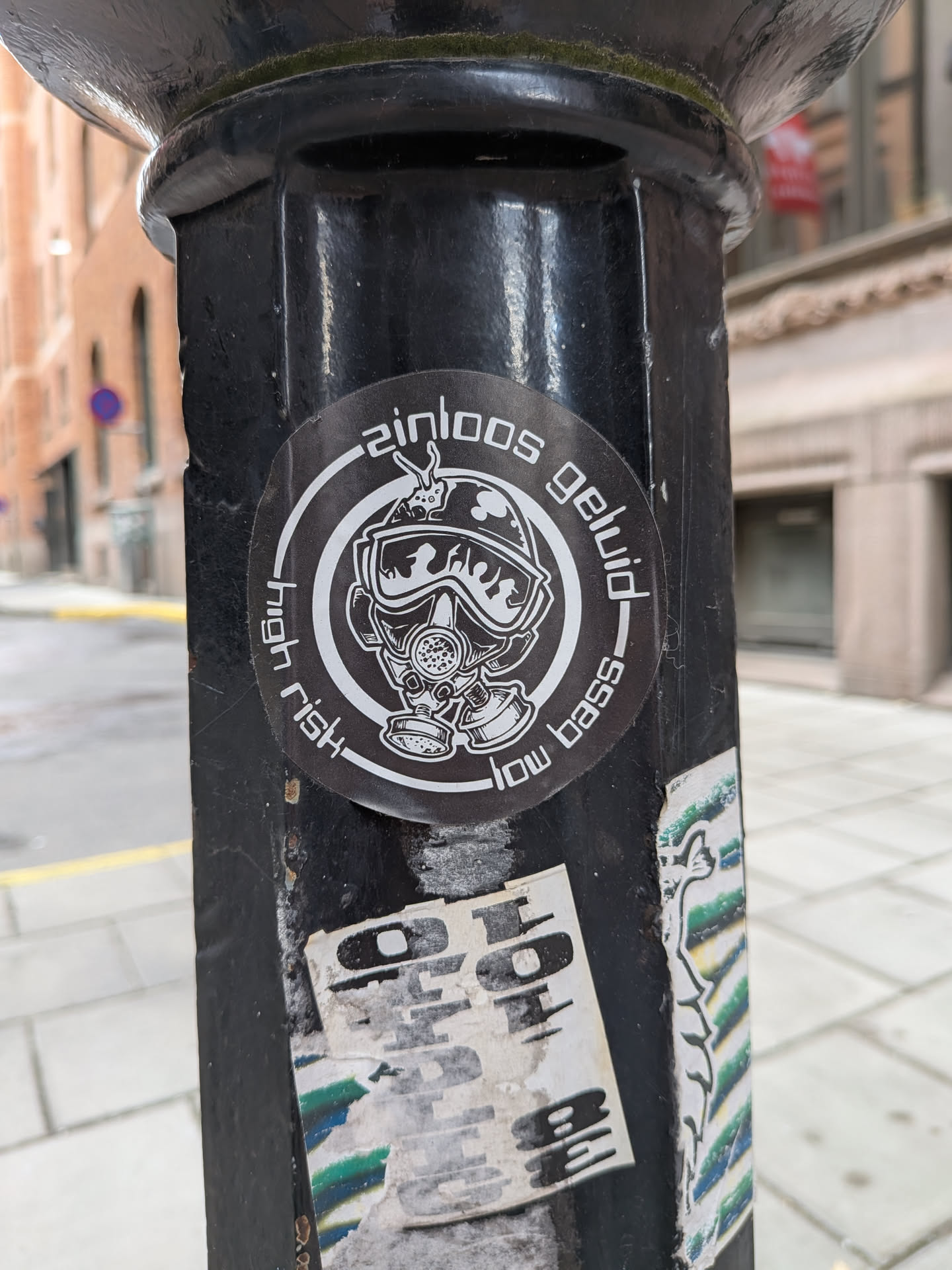 A circular black and white sticker depicting a character in a gas mask and helmet, surrounded by a circular border containing the text 'zinloos geluid', 'high risk', and 'low bass'.