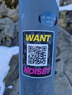 A sticker with a QR code and the text "WANT NOISE?" in yellow and pink. The background is black, and there are small smiley faces around the text and QR code.