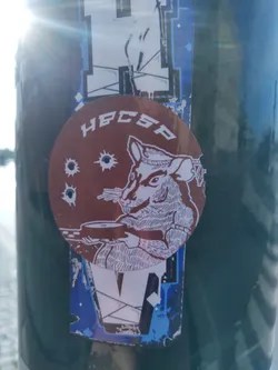 Street sticker A round brown sticker with a drawing of a rat holding what looks like a weapon. There are three bullet holes visible on the sticker around the rat. The text HBCSP is written on top of the sticker. The sticker is placed on a dark green surface, with other stickers underneath and nearby. 