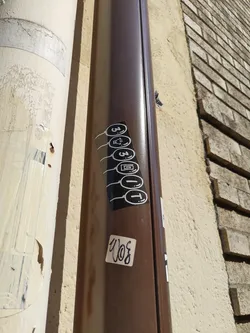 A vertical sticker featuring five circular icons connected by lines. The icons appear to be stylized symbols, possibly representing different functions or concepts. The sticker is affixed to a brown downspout on a building.  Another smaller sticker with illegible writing is below it.