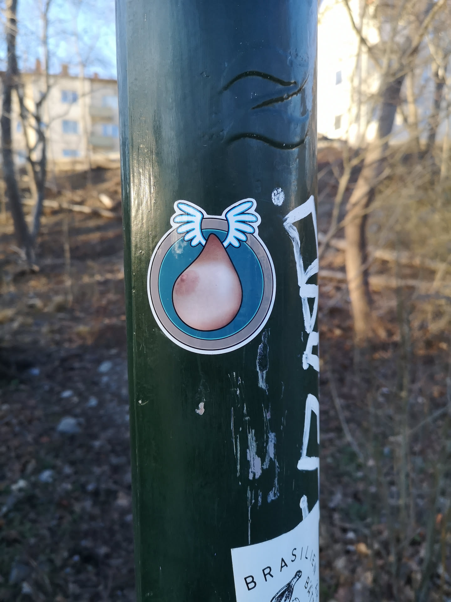 The sticker is predominantly circular, featuring a stylized human breast depicted in a teardrop shape. The breast is rendered in a light skin tone with a distinct brown nipple. From the top of the breast, a pair of small, light blue wings with white outlines extend upwards. The central image is set against a teal-blue circular background, which is framed by a thin grey border and a wider white outer border, giving the entire sticker a clean, cut-out appearance.