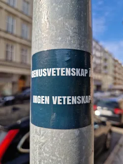 A dark teal rectangular sticker affixed to a lamppost. The text on the sticker is in Swedish and reads: "Genusvetenskap är ingen vetenskap" which translates roughly to "Gender studies are not science".