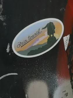 Street sticker An oval sticker with a white border showing a headless statue silhouette on a pedestal pointing left. The background features a gradient sunset sky over hills, with the cursive text 'Ruin tourist' written on the left.