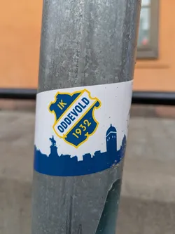 This is a sticker of IK Oddevold, a Swedish football club, fixed on a metallic pole. The sticker features the club's crest in blue and yellow, including the founding year "1932". Below the crest is a blue silhouette of buildings and monuments.