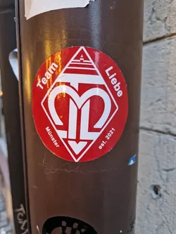 Red and white diamond-shaped sticker with a logo and text. The logo is a stylized "M" with an arrow pointing down. The text around the logo says "Team Liebe" at the top and "Münster est. 2021" at the bottom.