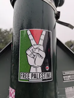 Street sticker The sticker is rectangular with a prominent white border. Its background is composed of three vertical color blocks: green on the left, red in the top right, and black in the bottom right, mirroring the colors of the Palestinian flag. A large, stylized white hand with black outlines is centered, making a peace sign (V-sign) with its index and middle fingers extended upwards. Below the hand, a white rectangular banner spans the width of the sticker, featuring the bold black text "FREE PALESTINE". In the bottom right corner of the main image area, above the text banner, there is a small circular symbol depicting a red and black flag, which is the Antifascist Action (Antifa) emblem. The sticker is visibly wet with numerous water droplets on its surface.