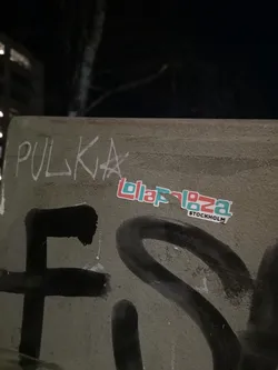 Street sticker A rectangular sticker featuring the word 'Lollapalooza' in a distinctive, blocky font. The letters are horizontally split, with the upper half in a light teal/blue and the lower half in a reddish-orange/coral. A thin white outline surrounds the text and the sticker's edge. Below 'Lollapalooza', the word 'STOCKHOLM' is written in a smaller, black sans-serif font, also with a white outline. The sticker is applied to a textured, grey-green surface, possibly concrete, which also bears other graffiti.