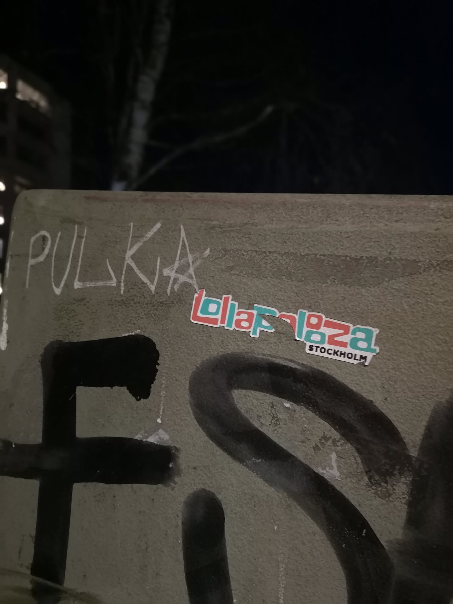 A rectangular sticker featuring the word 'Lollapalooza' in a distinctive, blocky font. The letters are horizontally split, with the upper half in a light teal/blue and the lower half in a reddish-orange/coral. A thin white outline surrounds the text and the sticker's edge. Below 'Lollapalooza', the word 'STOCKHOLM' is written in a smaller, black sans-serif font, also with a white outline. The sticker is applied to a textured, grey-green surface, possibly concrete, which also bears other graffiti.