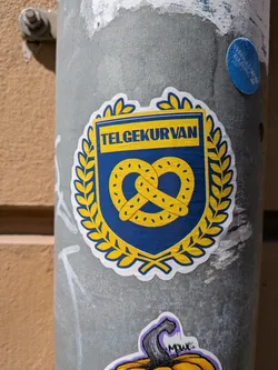 Street sticker Shield-shaped sticker in blue and yellow. Features a central yellow pretzel, a surrounding laurel wreath, and the text 'TELGEKURVAN' in a yellow banner at the top. Outlined with a white border.