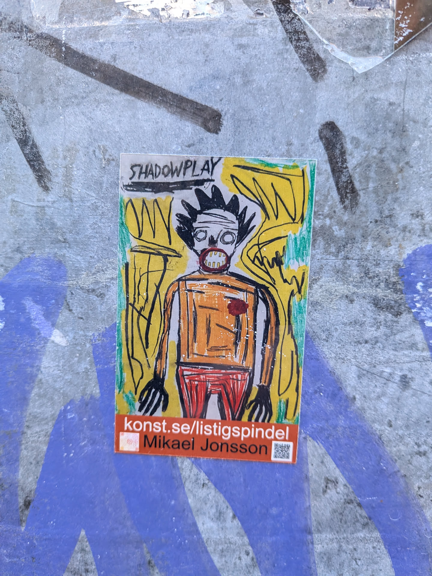 A rectangular sticker featuring a stylized, primitive human-like figure with a spiky black crown-like hairstyle. The figure has a bandaged head, wide-open eyes, and an open mouth revealing teeth, all outlined in black. Its body is orange with a red heart-like shape on the chest, and it wears red pants. The background is a vibrant yellow with energetic black and green scribbled lines, giving it a raw, expressive, and neo-expressionist feel. The top of the sticker reads 'SHADOWPLAY', and the bottom features a red bar with 'konst.se/listigspindel' and 'Mikael Jonsson', alongside a small, unidentifiable icon and a QR code.