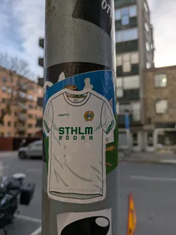 Street sticker A sticker depicting the jersey of Stockholm Södra FF, a Swedish football team. The sticker is white with green accents and the team's logo and name are clearly visible. The background of the sticker is light blue, providing a nice contrast to the jersey.