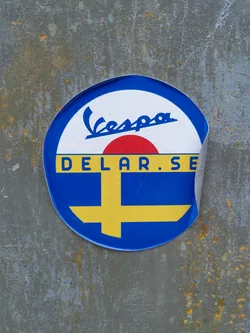 Street sticker A round sticker featuring the Vespa logo and the text "Delar.se" is affixed to a rusty gray metal surface. The sticker's design incorporates blue, yellow, red, and white colors, with the yellow and blue resembling the Swedish flag. The sticker shows some signs of peeling at the edge.