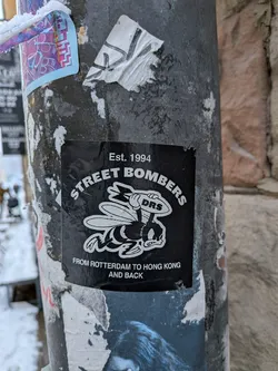 Street sticker A square black sticker featuring a white cartoonish bee with an angry expression, holding a bomb labeled 'DRS'. The text 'Est. 1994' is at the top, 'STREET BOMBERS' arcs above the bee, and 'FROM ROTTERDAM TO HONG KONG AND BACK' is at the bottom, all in white.