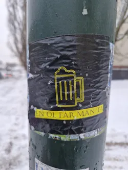 Street sticker A black and yellow sticker featuring a beer mug graphic and the text "En öl får man ta!" which translates to "One beer you can take!" in Swedish. The sticker is affixed to a lamppost in a snowy setting.