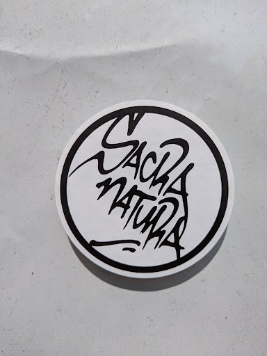 A circular sticker features a stylized black and white design. The words "Sack Natura" are rendered in a unique, almost graffiti-like font, positioned within the circle. The sticker is placed on a neutral, slightly textured surface.