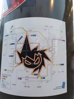 Street sticker A black and orange stylized graphic sticker is placed over a public transport map. The sticker features a jagged, starburst-like design with a face-like element in the center. The map appears to be of a tram or train system, with various lines and station names visible. The bottom right of the map features text in Dutch indicating the location, which seems to be Bougeniers plein.