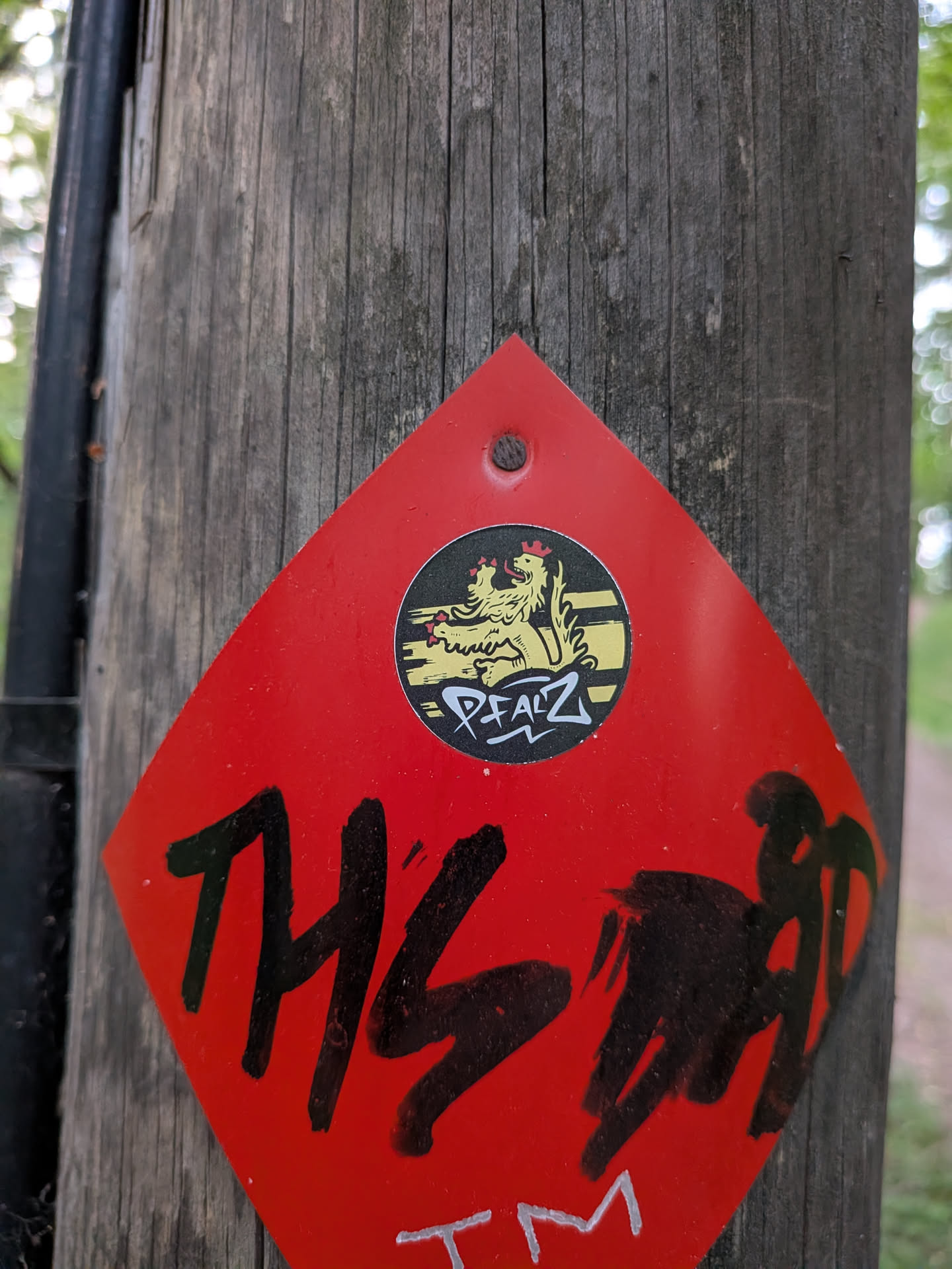 A circular black sticker featuring a golden Palatinate lion with a red crown, set against gold horizontal stripes. The word 'PFALZ' is written below in a white graffiti-style font. The sticker is placed on a red diamond-shaped sign.