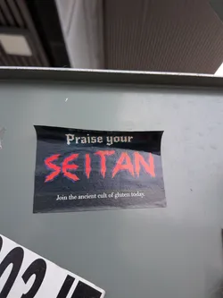 Street sticker The sticker is rectangular with slightly rounded top corners, set against a dark, possibly black or very dark blue, background. The text 'Praise your' is at the top in a white, gothic-style font. Below it, the word 'SEITAN' is prominently displayed in a large, distressed, brush-stroke style font, colored bright red with a subtle dark blue or purple outline/shadow, giving it a bold, almost aggressive look. At the bottom, in a smaller, white, serif font, it reads 'Join the ancient cult of gluten today.' The sticker combines a dark, somewhat ominous aesthetic with a humorous, plant-based food theme.