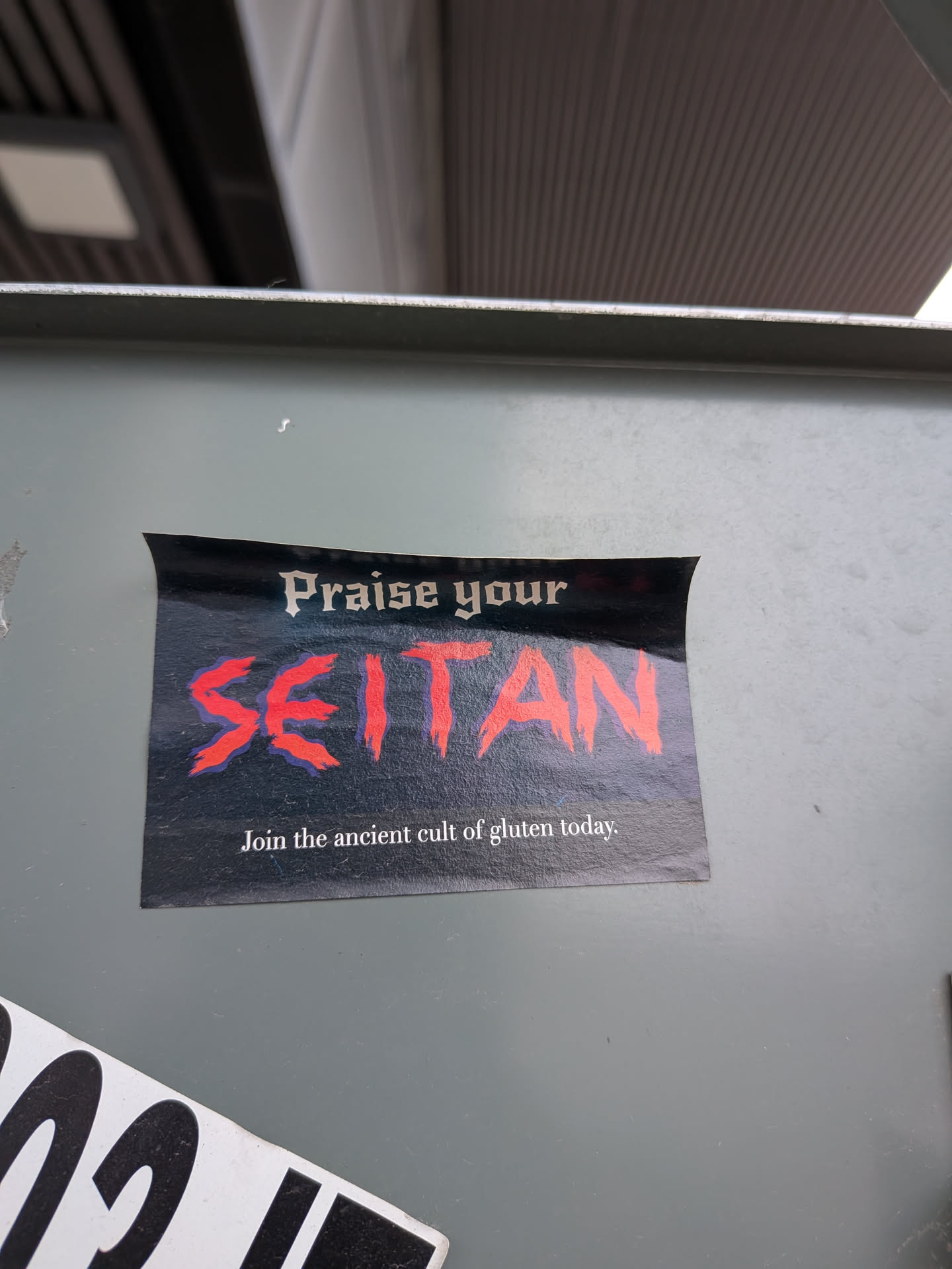 The sticker is rectangular with slightly rounded top corners, set against a dark, possibly black or very dark blue, background. The text 'Praise your' is at the top in a white, gothic-style font. Below it, the word 'SEITAN' is prominently displayed in a large, distressed, brush-stroke style font, colored bright red with a subtle dark blue or purple outline/shadow, giving it a bold, almost aggressive look. At the bottom, in a smaller, white, serif font, it reads 'Join the ancient cult of gluten today.' The sticker combines a dark, somewhat ominous aesthetic with a humorous, plant-based food theme.